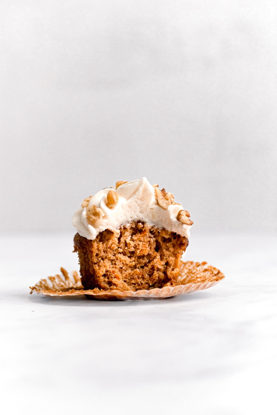 single frosted carrot cake cupcake with a bite taken out on a white surface