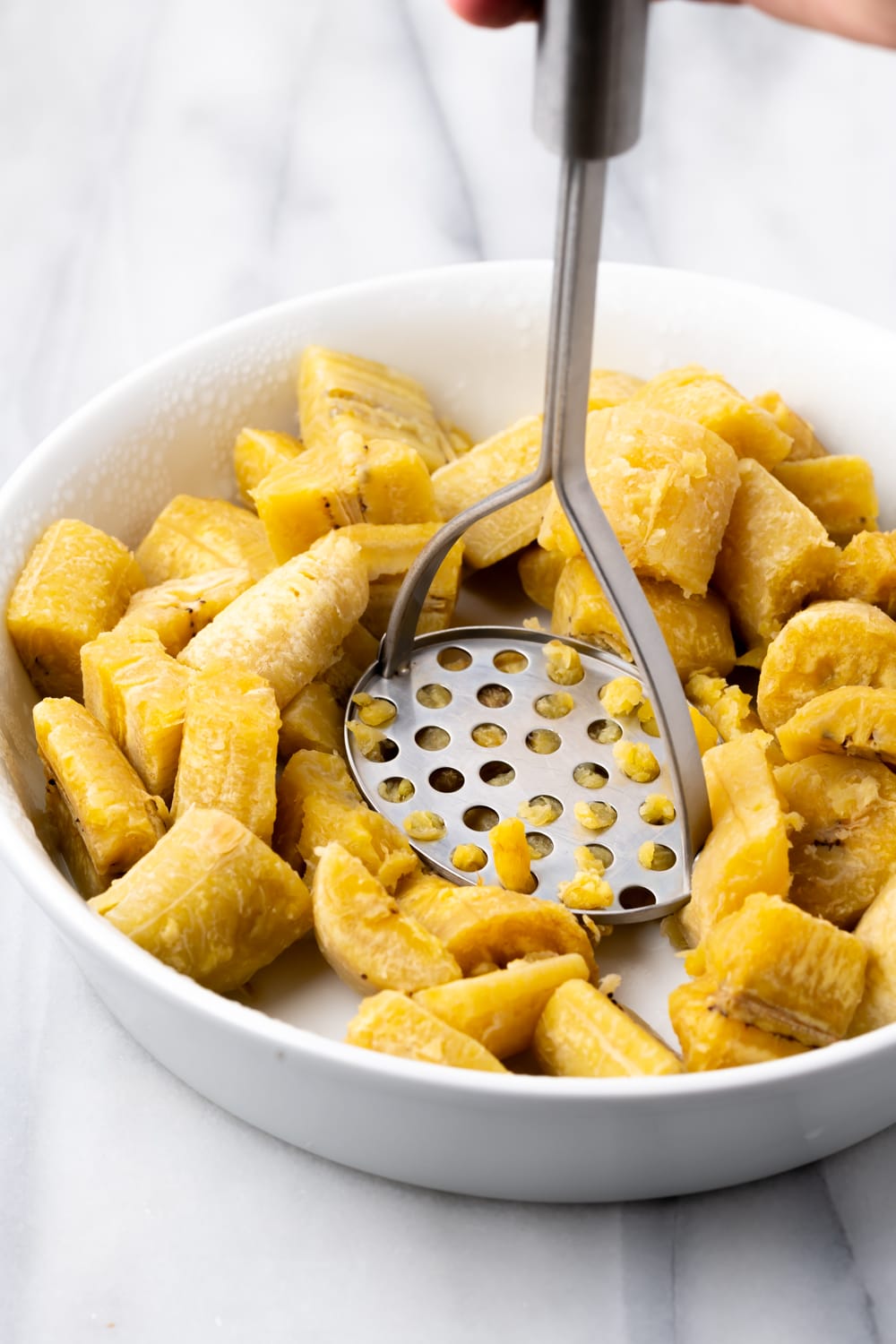 A potato masher crushing the boiled plantains in a large white bowl to make fufu de platano.