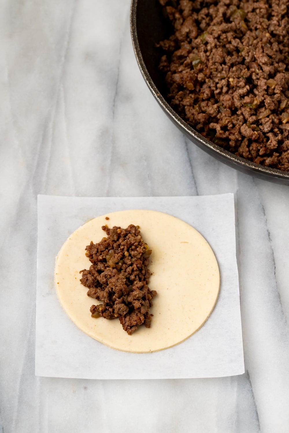 A round empanada dough with half filled with a beef filling and the pan of beef filling on the side