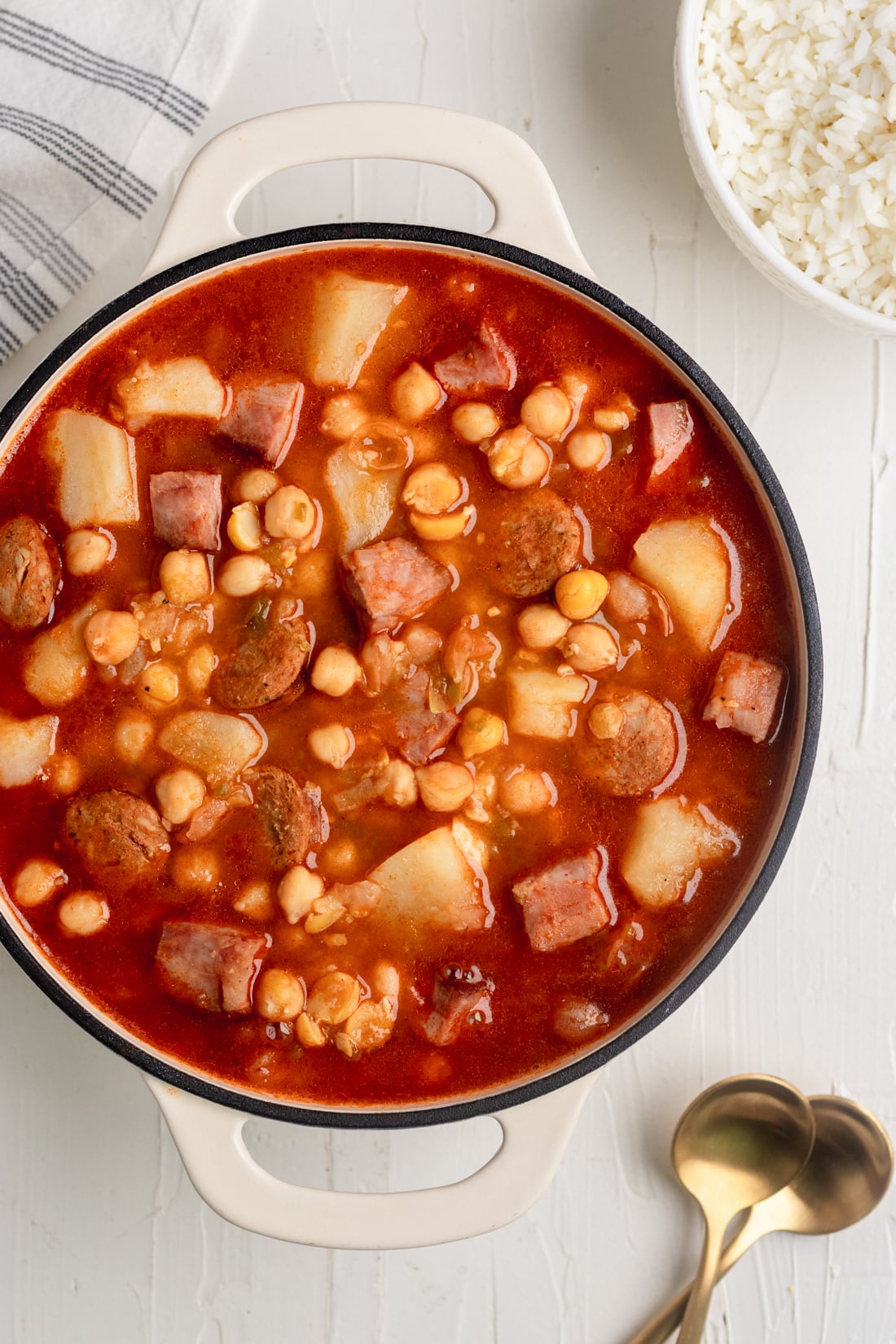 cuban chickpea stew in a dutch oven with 2 gold spoons and a bowl of rice on a white table