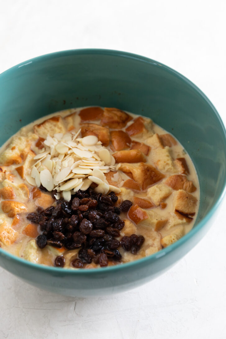 Creamy bread pudding with raisins, almonds, and custard in a teal bowl.